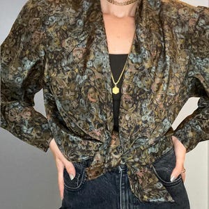 May include: A vintage floral blouse in brown, green, and pink hues, knotted at the waist. The long-sleeved top is worn with a black under-top and dark denim jeans. A gold necklace and choker are also visible.