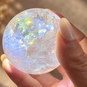 May include: A clear, spherical crystal ball featuring internal fractures that create a multi-colored iridescent effect. The surface is smooth and reflective, catching light to display flashes of blue, yellow, and white throughout the sphere.