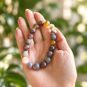 May include: A stretch bracelet composed of smooth, spherical beads in various shades of brown, gray, white, and yellow. The beads feature natural banded patterns and translucent qualities, arranged in a circular formation on a flexible cord.