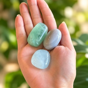 May include: Three smooth, polished tumbled stones in shades of light green, soft gray, and pale blue. Each stone has a rounded, organic shape with a glossy, reflective surface that catches the light, showcasing natural variations in color and texture.