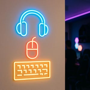 May include: Neon sign featuring a blue headset, a red mouse, and an orange keyboard. The gaming-themed sign is mounted on a neutral-colored wall, with a glimpse of a gaming room in the background.