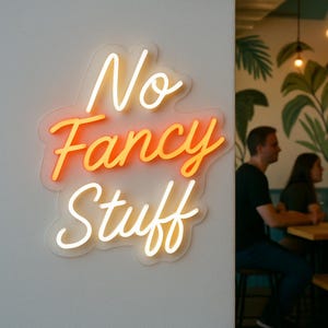 May include: A neon sign with the words "No Fancy Stuff" in a cursive font. The word "Fancy" is in orange, while "No" and "Stuff" are in white. The sign is mounted on a white wall. In the background, two people are seated at a table.