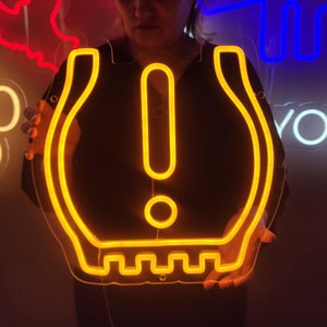 Puede incluir: Un letrero de neón con forma de signo de exclamación dentro de un círculo. El letrero es amarillo y lo sostiene una persona.