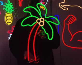 Könnte beinhalten: Eine Person hält ein leuchtend grünes Neon-Palmenzeichen mit roten Stielen. Das Schild leuchtet und im Hintergrund befinden sich weitere Neon-Schilder, darunter ein Ananas, ein Hund, ein Reh und die Worte "Say Cheeze!"