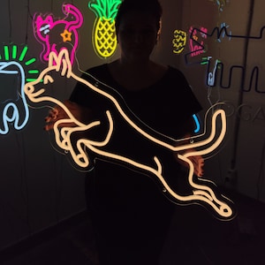 May include: A person holding a neon sign of a dog jumping in the air. The sign is white and the background is black. The person is wearing a black shirt. There are other neon signs in the background, including a pineapple, a cat, a tooth, and the word "DONUTS".