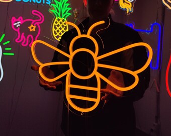 May include: A neon sign shaped like a bee with a yellow outline. The bee is lit up and glowing brightly. The sign is held by a person in front of a wall with other neon signs.