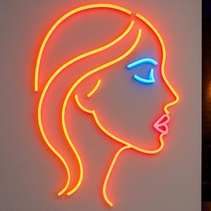 May include: Neon sign depicting a woman's profile with orange and yellow outlines. The closed eye is blue, and the lips are pink. The sign is mounted on a light gray wall, with a blurred background of a bar.