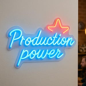 Könnte beinhalten: Ein Neonschild mit den Worten "Production power" in blauer Schrift und einem roten Stern darüber. Das Schild ist an einer weißen Wand angebracht. Das Schild ist ein dekorativer Gegenstand.