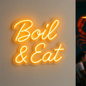 May include: A neon sign in a cursive font, illuminated with a warm orange glow, displaying the words "Boil & Eat". The sign is fixed to a light-coloured wall. In the background, a restaurant scene with a lobster mural is visible.
