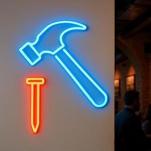 May include: A neon sign featuring a blue hammer and a red and yellow nail. The hammer is angled, and the nail is positioned below it. The sign is mounted on a light gray wall.