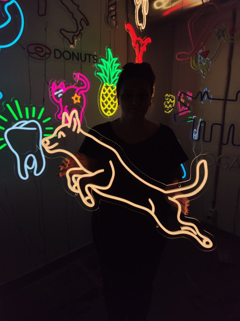 May include: A person holding a neon sign of a dog jumping in the air. The sign is white and the background is black. The person is wearing a black shirt. There are other neon signs in the background, including a pineapple, a cat, a tooth, and the word "DONUTS".
