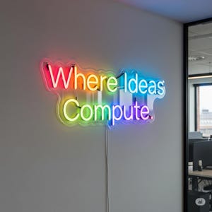 May include: A neon sign on a wall displays the text "Where Ideas Compute" in a rainbow of colors. The sign is mounted on a light gray wall with a clear plastic backing. A power cord hangs below the sign.