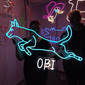 May include: Two people pose with a neon blue sign that says "OBI" and a neon blue dog design. The dog is holding a neon blue alligator in its mouth. The sign is hanging from the ceiling.