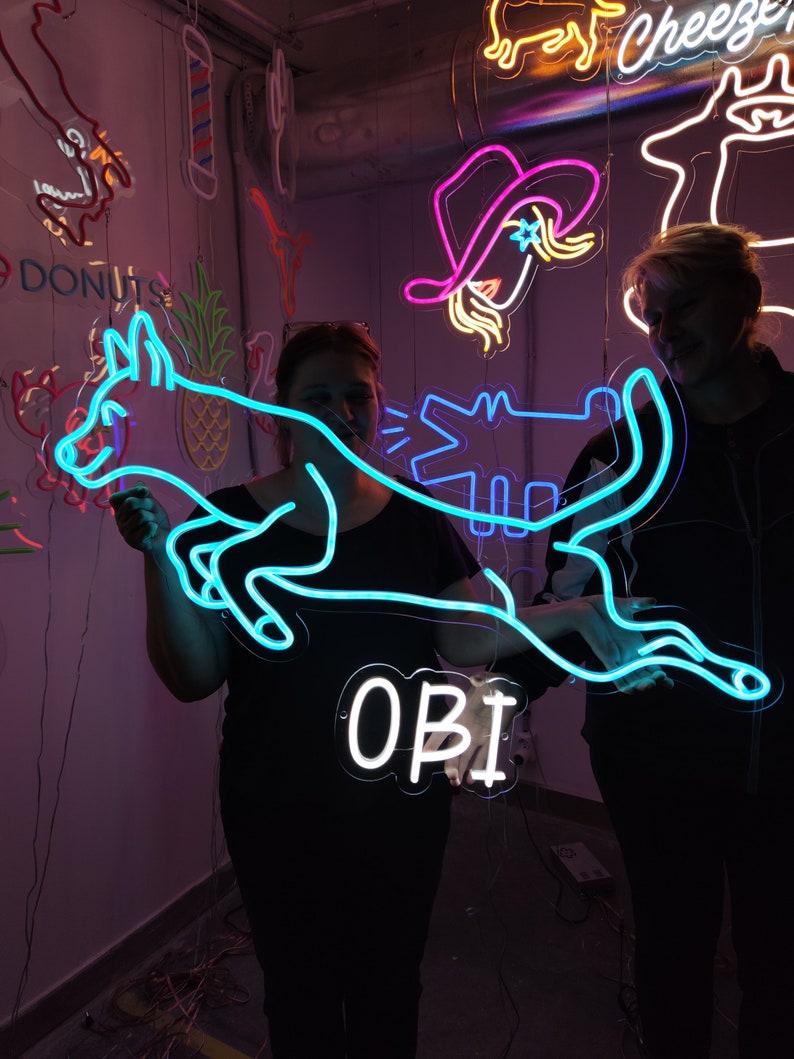 May include: Two people pose with a neon blue sign that says "OBI" and a neon blue dog design. The dog is holding a neon blue alligator in its mouth. The sign is hanging from the ceiling.
