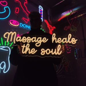 May include: A person holding a white neon sign that reads "Massage heals the soul". The sign is lit up and the person is standing in front of a wall with other neon signs.