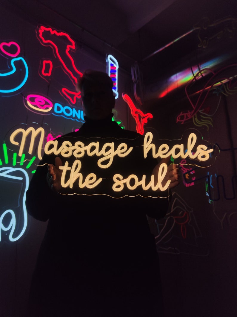 May include: A person holding a white neon sign that reads "Massage heals the soul". The sign is lit up and the person is standing in front of a wall with other neon signs.