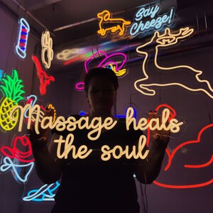 May include: A person holding a neon sign that says "Massage heals the soul". The sign is white with a yellow glow. There are other neon signs in the background, including a deer, a dog, and a cowboy hat.