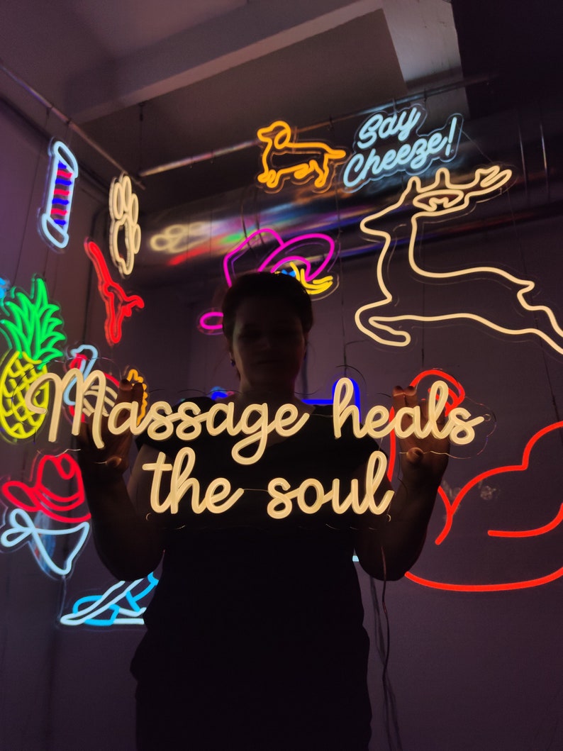 May include: A person holding a neon sign that says "Massage heals the soul". The sign is white with a yellow glow. There are other neon signs in the background, including a deer, a dog, and a cowboy hat.