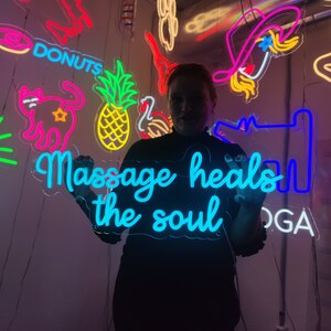 May include: A person holding a blue neon sign that says "Massage heals the soul". The sign is surrounded by other neon signs with various designs, including a pineapple, a cat, and a donut.