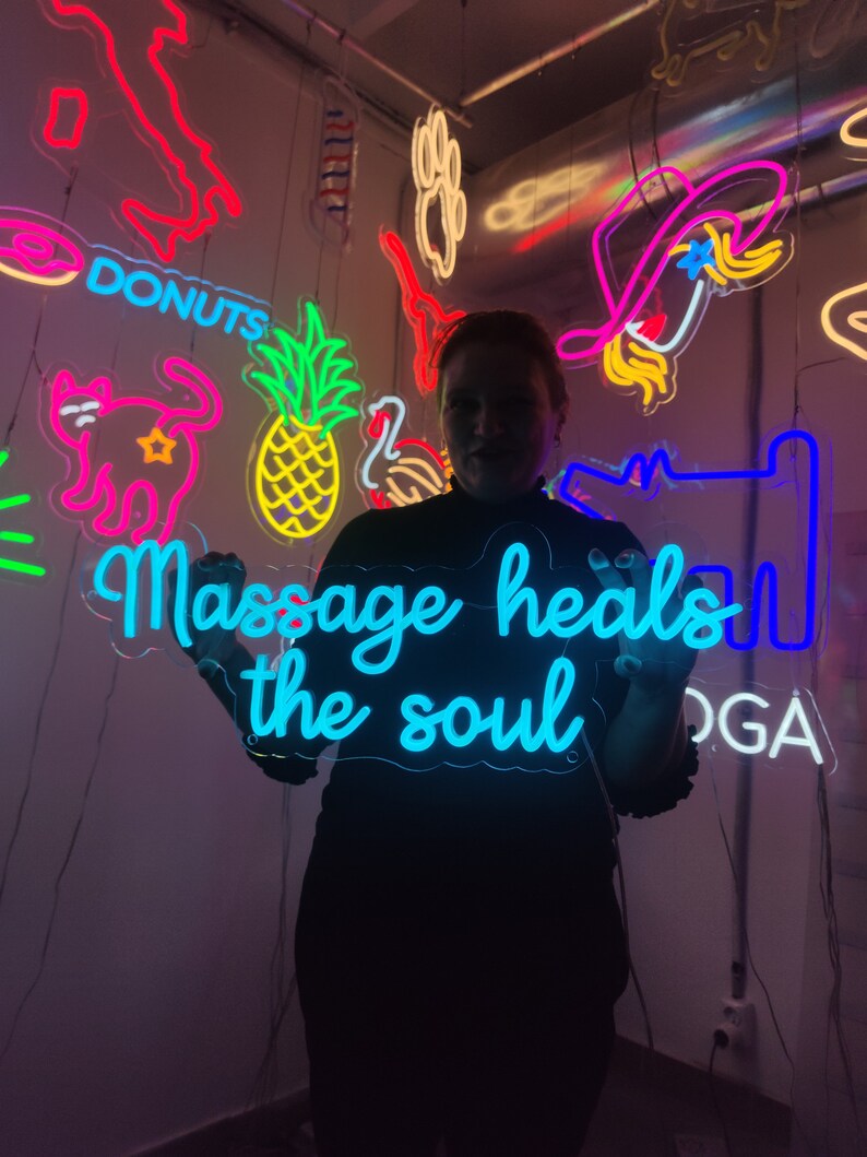 May include: A person holding a blue neon sign that says "Massage heals the soul". The sign is surrounded by other neon signs with various designs, including a pineapple, a cat, and a donut.