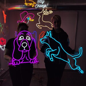 May include: Two people are holding neon signs. The sign on the left is purple and depicts a basset hound. The sign on the right is blue and depicts a dog jumping. The text "Say Cheeze!" is visible in the background.