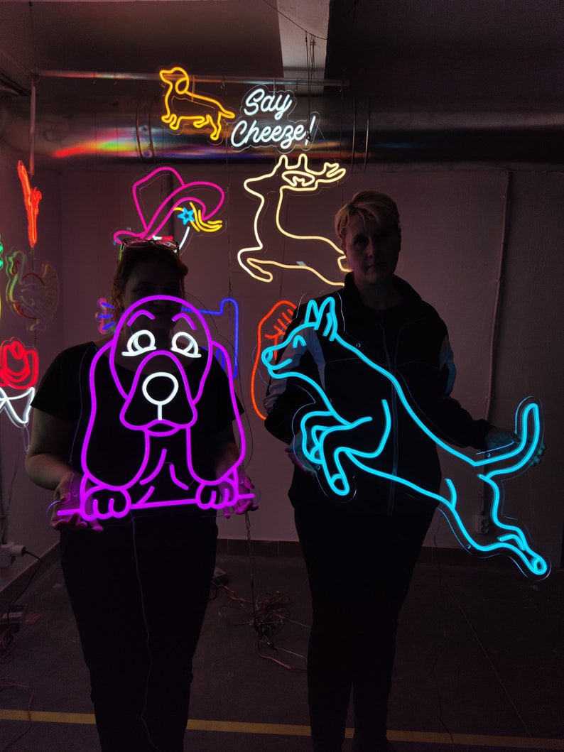 May include: Two people are holding neon signs. The sign on the left is purple and depicts a basset hound. The sign on the right is blue and depicts a dog jumping. The text "Say Cheeze!" is visible in the background.