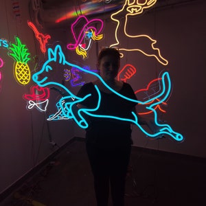 May include: A person stands in front of a wall with neon signs. The signs are in various shapes and colors, including a blue dog, a red deer, a yellow pineapple, and a pink cowboy hat. The text "Say Cheeze!" is on one of the signs.