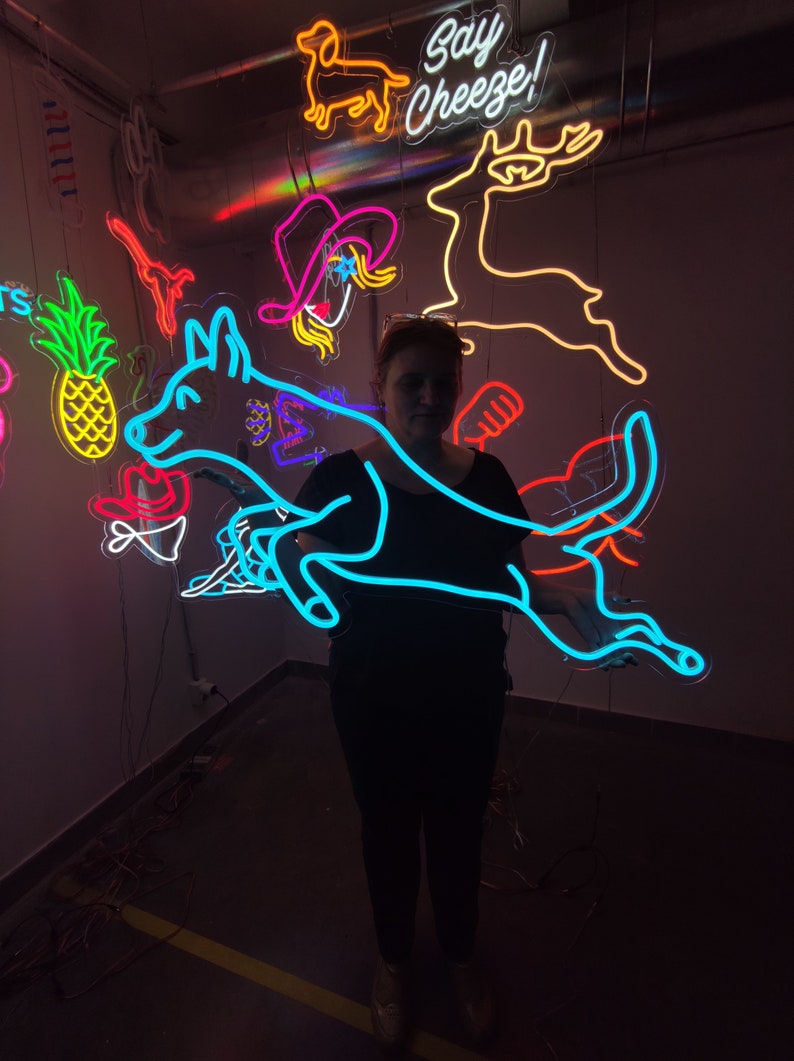 May include: A person stands in front of a wall with neon signs. The signs are in various shapes and colors, including a blue dog, a red deer, a yellow pineapple, and a pink cowboy hat. The text "Say Cheeze!" is on one of the signs.