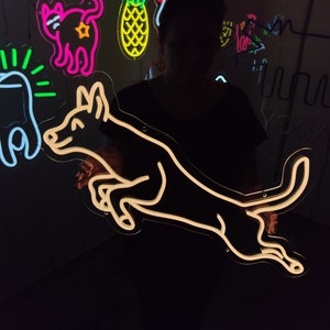 May include: A white neon sign of a dog jumping in the air. The sign is lit up and is hanging on a wall. There are other neon signs in the background, including a cat, a pineapple, and a bird.