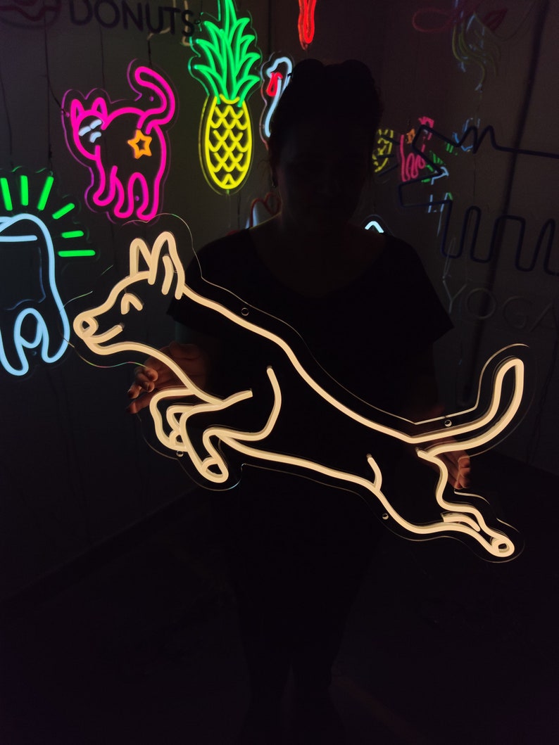 May include: A white neon sign of a dog jumping in the air. The sign is lit up and is hanging on a wall. There are other neon signs in the background, including a cat, a pineapple, and a bird.