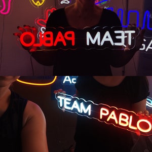 May include: Two neon signs with the text "TEAM PABLO" in white and red. The signs are held by two people.
