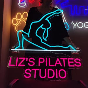 Może przedstawiać: Neonowy znak z niebieskim sylwetowym wizerunkiem osoby wykonującej pozycję jogi. Na znaku widnieje napis "Liz's Pilates Studio" w różowym neonie.