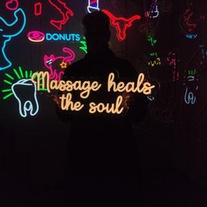 May include: A neon sign with the text "Massage heals the soul" in a yellow-orange color. The sign is held by a person in front of a wall with other neon signs, including a donut, a cat, a tooth, and a barber pole.