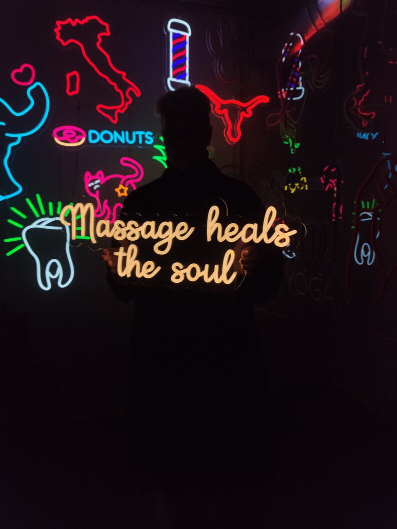 May include: A neon sign with the text "Massage heals the soul" in a yellow-orange color. The sign is held by a person in front of a wall with other neon signs, including a donut, a cat, a tooth, and a barber pole.