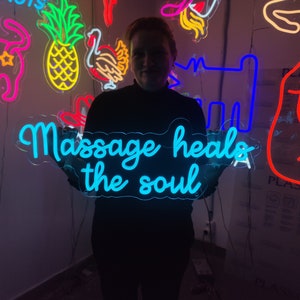 May include: A person holding a blue neon sign that says "Massage heals the soul". The sign is surrounded by other neon signs in various colors and shapes.