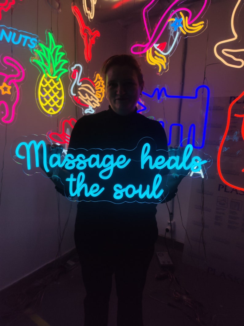 May include: A person holding a blue neon sign that says "Massage heals the soul". The sign is surrounded by other neon signs in various colors and shapes.