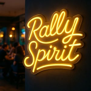 May include: A neon sign with the words "Rally Spirit" in a yellow, cursive font. The sign is mounted on a dark gray wall, with a blurred background of a restaurant or bar.