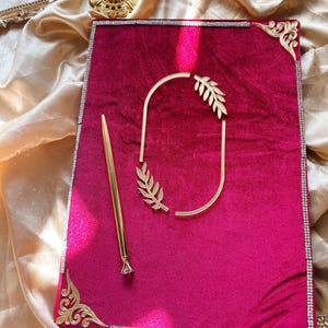 May include: A magenta velvet guest book with gold accents. The book has a gold pen with a diamond-shaped tip, a gold frame, and a gold decorative element. The edges of the book are lined with small, clear rhinestones.