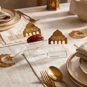 Könnte beinhalten: Eine Tischdekoration mit goldenen Elementen. Enthält eine Dattel in einem transparenten Acrylhalter, einen goldenen Löffel und eine Gabel sowie eine Mond- und Stern-Dekoration. Der Tisch ist mit einer weißen Tischdecke bedeckt.