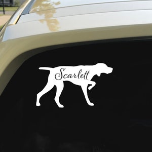 Puede incluir: Una calcomanía de vinilo blanco para coche con la silueta de un perro y el nombre "Scarlett" en cursiva. El perro está representado en posición de caminar. La pegatina está adherida a la ventana trasera de un coche.