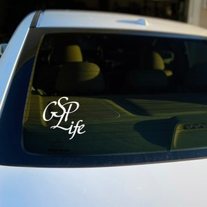 May include: White car rear window with a white decal that reads "GSP Life" in a cursive font. The decal is centered on the glass. The car is parked outside on a sunny day.