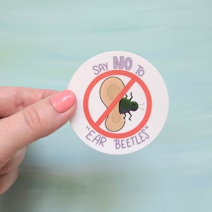 May include: A round sticker with the text "SAY NO TO 'EAR BEETLES'" in purple. The design features a cartoon ear with a green beetle, crossed out with a red line. The sticker is held by a hand.