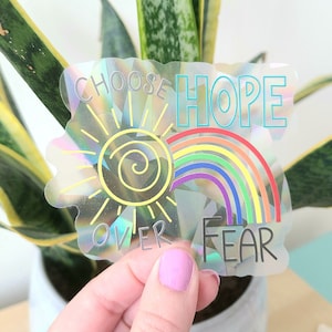 May include: A holographic sticker with a rainbow and sun design. The sticker says "Choose Hope Over Fear".