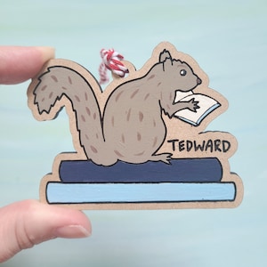 May include: Wooden ornament featuring a cartoon squirrel reading a book, perched atop two stacked books. The squirrel is painted in shades of brown and gray, holding a white book. The ornament has the name "TEDWARD" and a red and white string.
