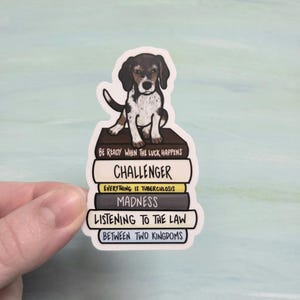 May include: A sticker featuring a beagle puppy sitting atop a stack of books. The books have titles like "Challenger" and "Madness." The sticker has a white border and is held by a hand against a light blue background.