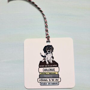 May include: A square white ornament featuring a black, brown, and white puppy sitting atop a stack of books. The books have titles like "Challenger" and "Madness." A red, green, and white string is attached for hanging.