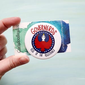 May include: A white button with a red, white, and blue design. The button features a cartoon eagle with a star on its head and the text "GOVERNERDS RISE OF THE RESISTANCE".