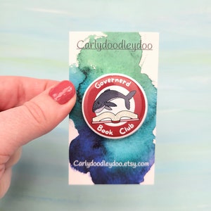 May include: A red and white button with a whale reading a book. The button says "Governerd Book Club".