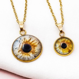 May include: Two gold-coloured chain necklaces. Each has a round pendant with a detailed eye design. The irises are a mix of blue, brown, and yellow, with a dark pupil. The pendants are set in gold-toned bezels.