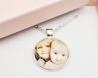 May include: A silver pendant necklace with a circular photo of two children inside a pink gift box. The necklace is displayed on a white surface with white roses in the background. The pendant is approximately 1 inch in diameter.